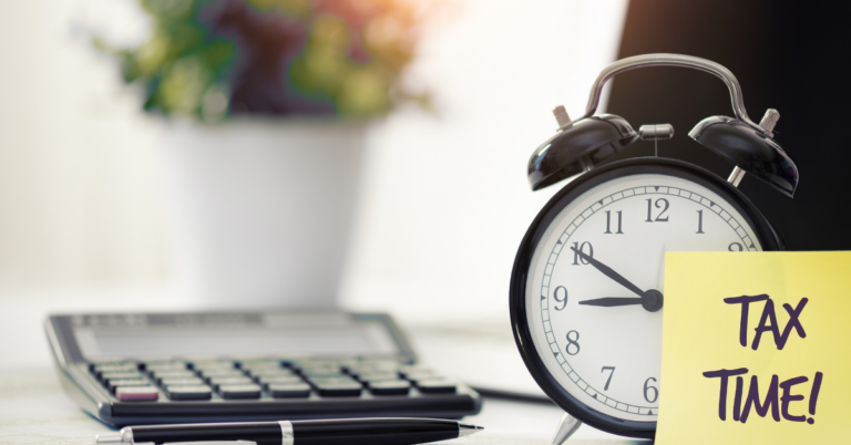 Desk with calculator, pen, alarm clock and a yellow post it note saying tax time